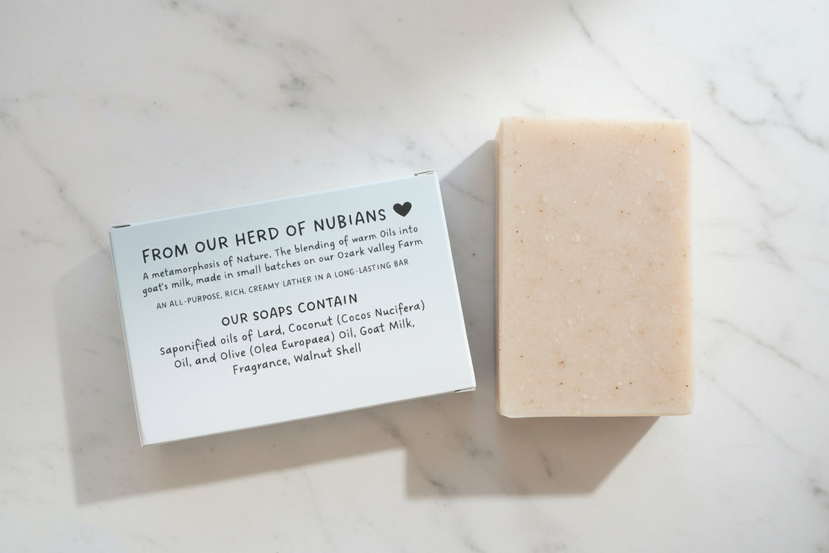 A rectangular bar of pale beige soap with small dark specks sits next to a white paper label card on a textured white towel. The label reads 'FROM OUR HERD OF NUBIANS' and lists ingredients including coconut oil, olive oil, goat milk, and walnut shell.