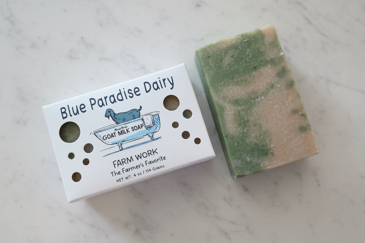 A handmade goat milk soap bar with green and cream swirls sits next to its white cardboard packaging box labeled 'Blue Paradise Dairy' on a textured white towel.