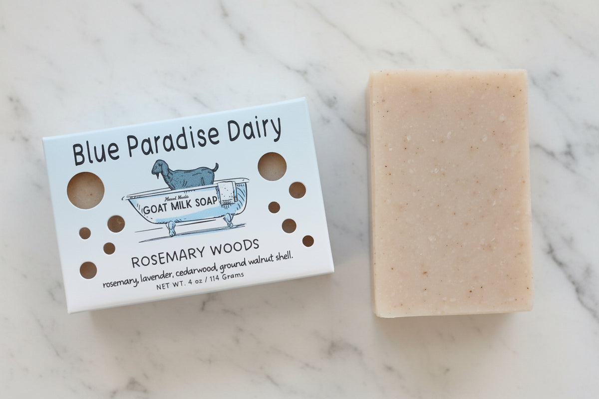 A bar of Blue Paradise Dairy Goat Milk Soap in the Rosemary Woods scent, shown next to its white paper packaging box on a textured beige fabric background.