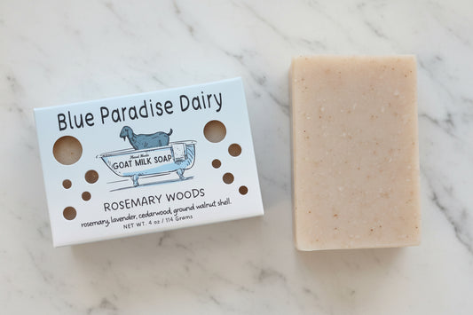 A bar of Blue Paradise Dairy Goat Milk Soap in the Rosemary Woods scent, shown next to its white paper packaging box on a textured beige fabric background.