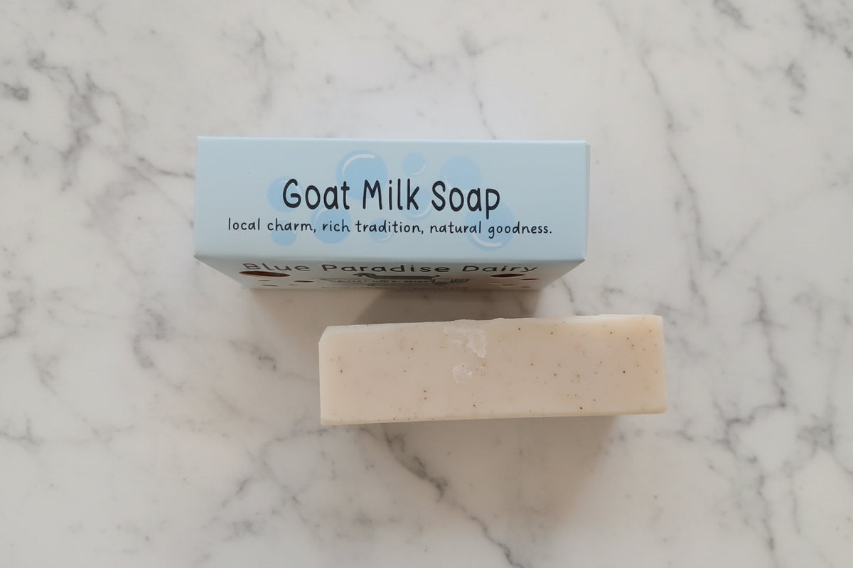 A bar of goat milk soap placed in front of its light blue packaging box on a textured beige towel. The box is labeled 'Goat Milk Soap' and 'Blue Paradise Dairy'.